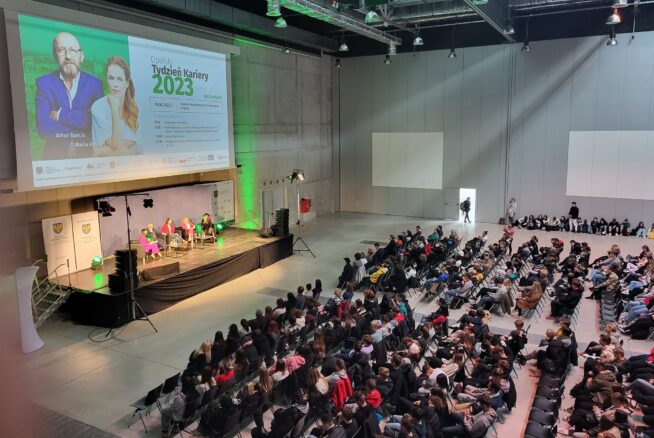 Duże wydarzenie w pomieszczeniu ze sceną, na której cztery osoby siedzą w dyskusji przed publicznością. Na ekranie wyświetlany jest program Týždeň Kaviarny 2023 ze zdjęciami dwóch prezenterów. Rzędy siedzących uczestników wypełniają salę, zwrócone w stronę sceny. Miejsce jest nowoczesne i przestronne.