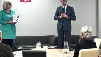 Mężczyzna w garniturze przemawia stojąc przed niewielką publicznością w sali konferencyjnej. Obok niego stoi kobieta trzymająca dokumenty. Na ścianie za nimi widnieje polskie godło. Ludzie siedzą przy stole, na którym stoją dzbanki z wodą i szklanki.
