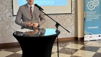 Mężczyzna w szarym garniturze przemawia na podium z mikrofonem w sali konferencyjnej. Za nim znajduje się ekran projektora wyświetlający slajd prezentacji w języku polskim i niebieski baner z kołami zębatymi, wskazujący na wydarzenie zawodowe lub edukacyjne.