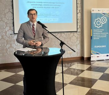 Mężczyzna w szarym garniturze przemawia na podium z mikrofonem w sali konferencyjnej. Za nim znajduje się ekran projektora wyświetlający slajd prezentacji w języku polskim i niebieski baner z kołami zębatymi, wskazujący na wydarzenie zawodowe lub edukacyjne.