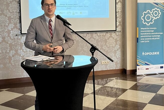Mężczyzna w szarym garniturze przemawia na podium z mikrofonem w sali konferencyjnej. Za nim znajduje się ekran projektora wyświetlający slajd prezentacji w języku polskim i niebieski baner z kołami zębatymi, wskazujący na wydarzenie zawodowe lub edukacyjne.