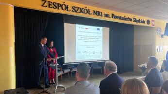 Dwóch prezenterów stoi na podium w szkolnej auli, twarzą do siedzących uczestników. Duży ekran projekcyjny za nimi wyświetla tekst. Na tle sceny widnieje napis Zespół Szkół Nr 1 im. Powstańców Śląskich. Pokój jest dobrze oświetlony i ma żółte ściany.