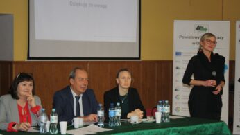 Cztery osoby siedzą przy zielonym stole z butelkami wody i papierami, zwrócone w stronę publiczności. Nieopodal stoi kobieta z mikrofonem. Za nimi znajduje się ekran projekcyjny wyświetlający 