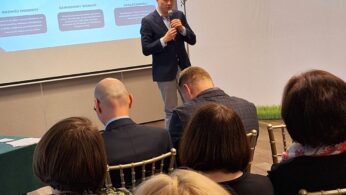 Mężczyzna w garniturze przemawia do mikrofonu przed siedzącą publicznością. Za nim duży ekran wyświetla tytuł 