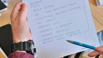 Osoba trzymająca papier z odręcznymi notatkami w języku polskim. Papier ma wydrukowany nagłówek dotyczący ścieżek edukacyjnych i potrzeb społeczności. W ręku znajduje się niebieski długopis. Inne materiały biurowe, notatniki i dokumenty są rozrzucone na drewnianym biurku w tle.