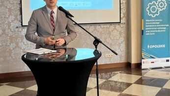 Mężczyzna w szarym garniturze i okularach stoi na czarnym podium z mikrofonem, przemawiając w pomieszczeniu. Za nim ekran projektora wyświetla prezentację w języku polskim. Po prawej stronie stoi kolorowy baner, a podłogę zdobią czarno-białe kafelki.