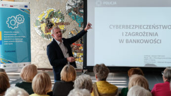Mężczyzna w garniturze stoi przed publicznością, gestykulując w kierunku ekranu wyświetlającego polską prezentację na temat cyberbezpieczeństwa i zagrożeń bankowych. Publiczność składa się głównie z osób starszych. W tle znajduje się kolorowy baner i mozaika ścienna.