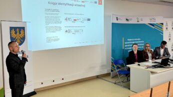 Mężczyzna mówi do mikrofonu obok ekranu wyświetlającego prezentację zatytułowaną Księga identyfikacji wizualnej. Cztery osoby siedzą przy biurku z laptopami. W pomieszczeniu widoczny jest niebieski baner i żółty emblemat heraldyczny.