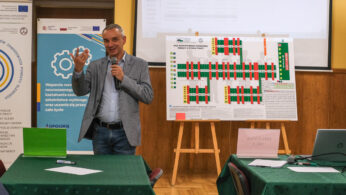 Mężczyzna w szarej marynarce mówi do mikrofonu, stojąc obok stołu z zielonymi obrusami. Za nim znajduje się tablica prezentacyjna z diagramami, baner z tekstem i logo oraz ekran. Dwa laptopy i papiery leżą na stołach na pierwszym planie.