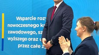 Mężczyzna w ciemnym garniturze i okularach stoi uśmiechnięty przed niebieskim tłem promocyjnym z polskim tekstem. Kobieta w niebieskiej kurtce, siedząca i klaszcząca, jest na pierwszym planie. Scena wygląda na oficjalne wydarzenie lub prezentację.