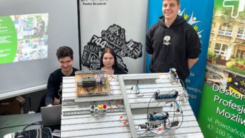Trzech uśmiechniętych młodych ludzi stoi i siedzi za dużym metalowym panelem przedstawiającym elektroniczny i pneumatyczny projekt. Na stole leżą laptopy, broszury i banery z tekstem i obrazami, co sugeruje targi naukowe lub wystawę technologiczną.