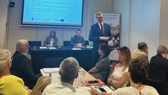 Mężczyzna w garniturze stoi i przemawia do grupy siedzącej przy stołach w sali konferencyjnej. Dwie osoby siedzą przy stole z laptopami przed ekranem prezentacji wyświetlającym tekst. Uczestnicy słuchają uważnie, niektórzy z notatnikami, pod jasnymi lampami sufitowymi.