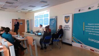 Mężczyzna w garniturze prezentuje z przodu sali konferencyjnej, używając mikrofonu i slajdów. Słuchacze siedzą przy biurkach z laptopami. Banery wyświetlają polski tekst o edukacji i regionie opolskim. Cztery osoby siedzą przy stole naprzeciwko publiczności.