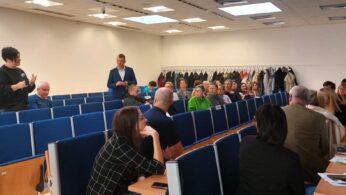 Grupa ludzi siedzi na niebieskich krzesłach w klasie lub sali seminaryjnej. Jedna osoba stoi po lewej stronie i mówi, podczas gdy inni uważnie słuchają. Płaszcze wiszą na wieszakach wzdłuż tylnej ściany. Papiery i laptopy leżą na biurkach na pierwszym planie.