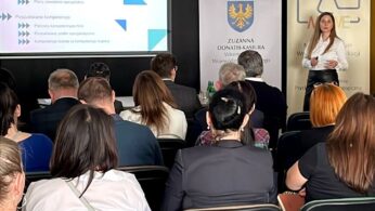 Kobieta stoi z przodu sali i przemawia do publiczności. Prezentuje w pobliżu ekranu projekcyjnego wyświetlającego tekst i grafikę w języku polskim. Uczestnicy siedzą przodem do niej, niektórzy robią notatki. Za prezenterką widoczne są banery i herb.