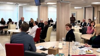 Grupa ludzi w strojach biznesowych siedzi przy okrągłych stołach w nowoczesnej sali konferencyjnej i uważnie słucha. Na stołach leżą papiery, książki i okulary. Duże okna wpuszczają światło dzienne. Niektóre osoby zwrócone są do przodu, podczas gdy inne odwracają się, by słuchać lub patrzeć w kamerę.