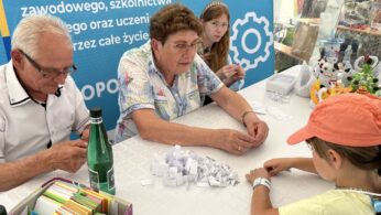 Dwoje starszych dorosłych i dwoje dzieci siedzi przy stole przykrytym papierowymi karteczkami, książkami i origami. W tle znajduje się niebieski baner z polskim tekstem. Scena wygląda na interaktywną aktywność edukacyjną lub rzemieślniczą podczas wydarzenia lub targów.