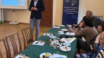 Mężczyzna stoi i przemawia przed ekranem projektora, wyświetlając prezentację dla czterech osób siedzących wokół zielonego stołu z papierami, notatnikami, filiżankami i talerzami. W tle znajduje się baner z ikonami naukowymi i rosyjskim tekstem. Pokój ma drewniane podłogi i krzesła.