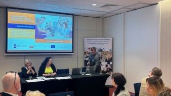 Grupa ludzi siedzi przy stole konferencyjnym w nowoczesnej sali konferencyjnej. Mężczyzna stoi w pobliżu ekranu projektora wyświetlającego prezentację. Uczestnicy zwróceni są twarzą do prelegenta, niektórzy robią notatki. Atmosfera jest profesjonalna i sprzyja współpracy.