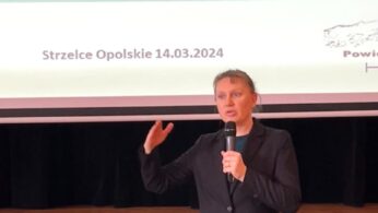 Kobieta w ciemnej marynarce i turkusowej spódnicy przemawia z uniesioną ręką. Za nią znajduje się ekran wyświetlający polski tekst, ikony osób wykonujących różne zawody i datę 14.03.2024. Sceneria wygląda na formalną prezentację lub seminarium.