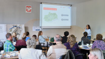 Kobieta stoi i przemawia do grupy siedzącej przy stołach w jasno oświetlonym pomieszczeniu. Mapa i diagramy są wyświetlane na ekranie za nią. Uczestnicy stoją twarzą do prezenterki, słuchając i patrząc na materiały. Na stołach widoczne są papiery, kubki i notatniki.