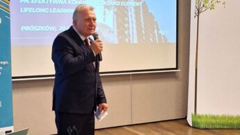 Mężczyzna w ciemnym garniturze i krawacie mówi do mikrofonu w sali konferencyjnej. Za nim wyświetlany jest slajd prezentacji z polskim tekstem i obrazem nowoczesnego budynku. Po prawej stronie znajduje się panel z ilustracją drzewa. Na pierwszym planie znajduje się zielony stół.