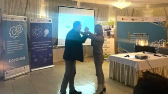 Mężczyzna i kobieta stoją naprzeciwko siebie, dotykając się rękami, jakby tańczyli, w sali konferencyjnej. Za nimi znajdują się banery z polskim tekstem, stół z projektorem, oświetlony żyrandol i prezentacja wyświetlana na ekranie. Pomieszczenie jest delikatnie oświetlone i udekorowane.