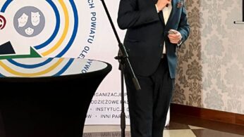 Mężczyzna w ciemnym garniturze stoi na podłodze w kratkę, trzymając mikrofon i przemawiając. Za nim znajduje się baner z polskim tekstem, logo i kolorową okrągłą grafiką. Na pierwszym planie widoczny jest stojący stół i stojak na mikrofon.