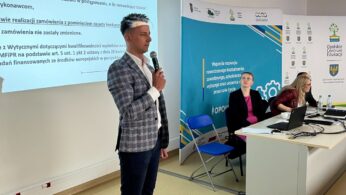 Mężczyzna w kraciastej marynarce i czarnych spodniach przemawia do mikrofonu w jasno oświetlonej sali konferencyjnej, podczas gdy dwie kobiety siedzą przy stole z laptopami. Na ścianie za nimi wyświetlany jest slajd prezentacji w języku polskim. W tle widoczne są banery i logo.