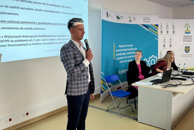 Mężczyzna w kraciastej marynarce i czarnych spodniach przemawia do mikrofonu w jasno oświetlonej sali konferencyjnej, podczas gdy dwie kobiety siedzą przy stole z laptopami. Na ścianie za nimi wyświetlany jest slajd prezentacji w języku polskim. W tle widoczne są banery i logo.