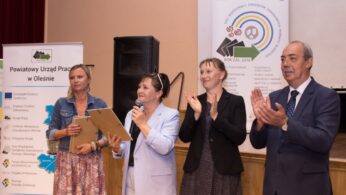 Cztery dorosłe osoby stoją przed banerami na imprezie wewnętrznej. Jedna kobieta w jasnej kurtce trzyma mikrofon i przemawia, podczas gdy inna kobieta obok niej trzyma tablicę. Dwie inne osoby - kobieta i mężczyzna w garniturze - stoją obok nich, uśmiechając się i klaszcząc.