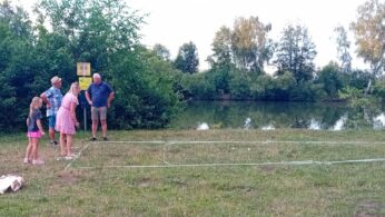 Cztery osoby stoją na trawie w pobliżu stawu, obok prostokątnej metalowej ramy na ziemi. Dwie dziewczynki i dwie osoby dorosłe wydają się ustawiać lub sprawdzać ramę. Drzewa i krzewy otaczają staw w tle, a w pobliżu znajduje się torba.