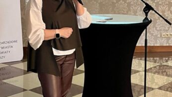 Kobieta z ciemnymi włosami spiętymi w kok, ubrana w białą koszulę i brązową kamizelkę, mówi do mikrofonu. Stoi obok wysokiego czarnego stołu. Za nią znajduje się ekran projekcyjny z tekstem i dekoracyjna ściana. Podłoga ma wzór w kratkę.