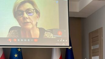 Kobieta w krótkich blond włosach i okularach rozmawia podczas rozmowy wideo wyświetlanej na ekranie projektora. Pod ekranem widoczne są dwie polskie flagi i dwie flagi Unii Europejskiej. Pokój ma beżowe ściany i zamknięte drewniane drzwi.