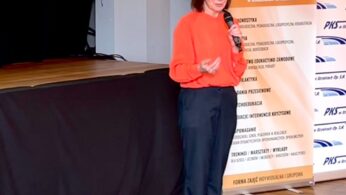 Kobieta o prostych brązowych włosach, ubrana w jasnopomarańczową bluzkę i ciemne spodnie, stoi na drewnianej podłodze trzymając mikrofon. Przemawia podczas wydarzenia w pomieszczeniu obok ekranu i banerów informacyjnych napisanych po polsku.