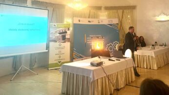 Mężczyzna w garniturze mówi do mikrofonu w jasnej sali konferencyjnej. Jest tam ekran projekcyjny pokazujący niebieski slajd z polskim tekstem oraz kilka stołów z białymi obrusami, dzbanki z kawą i ludzie siedzący, słuchający. W tle widoczne są banery i żyrandol.