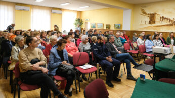 Duża grupa dorosłych, głównie seniorów, siedzi uważnie w rzędach krzeseł w dobrze oświetlonym pokoju. Niektórzy rozmawiają cicho, podczas gdy inni zwróceni są twarzami do przodu. Projektor i pokryte zielenią stoły znajdują się z przodu; oprawione obrazy zdobią żółte ściany.