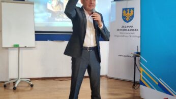 Mężczyzna w szarym garniturze i jasnoniebieskich butach stoi na drewnianej podłodze, trzymając mikrofon i gestykulując w górę. Za nim znajduje się ekran wyświetlający polski tekst i obrazy, tablica i banery z oficjalnymi logo. Wygląda to na formalną prezentację lub wykład.