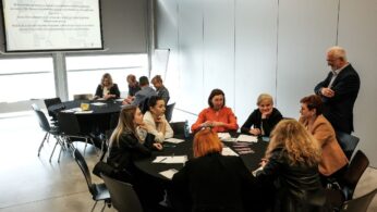 Ludzie siedzą przy okrągłych stołach w nowoczesnej sali konferencyjnej, zaangażowani w dyskusje grupowe. Na stołach leżą papiery i długopisy. Mężczyzna stoi i rozmawia z grupą kobiet. Na ścianie z przodu widoczny jest wyświetlany slajd z polskim tekstem.