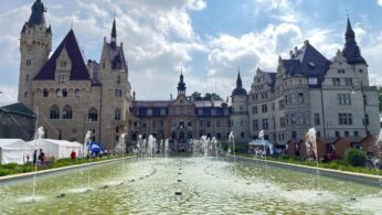 Duży kamienny zamek ze spiczastymi wieżami i stromymi dachami stoi za prostokątną fontanną z wieloma małymi strumieniami wody. Niebo jest częściowo zachmurzone, a ludzie spacerują wokół zamku, namioty widoczne po lewej stronie. Drzewa otaczają scenę w tle.