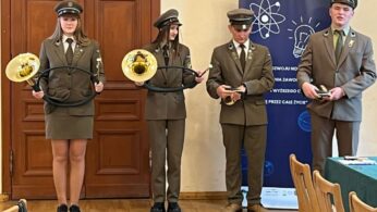 Czworo młodych ludzi w brązowych mundurach i czapkach stoi w pomieszczeniu. Dwie dziewczyny po lewej trzymają zakrzywione instrumenty dęte, a dwóch chłopców po prawej medale lub nagrody. Za nimi znajdują się drewniane drzwi i niebieski baner z grafiką o tematyce naukowej.
