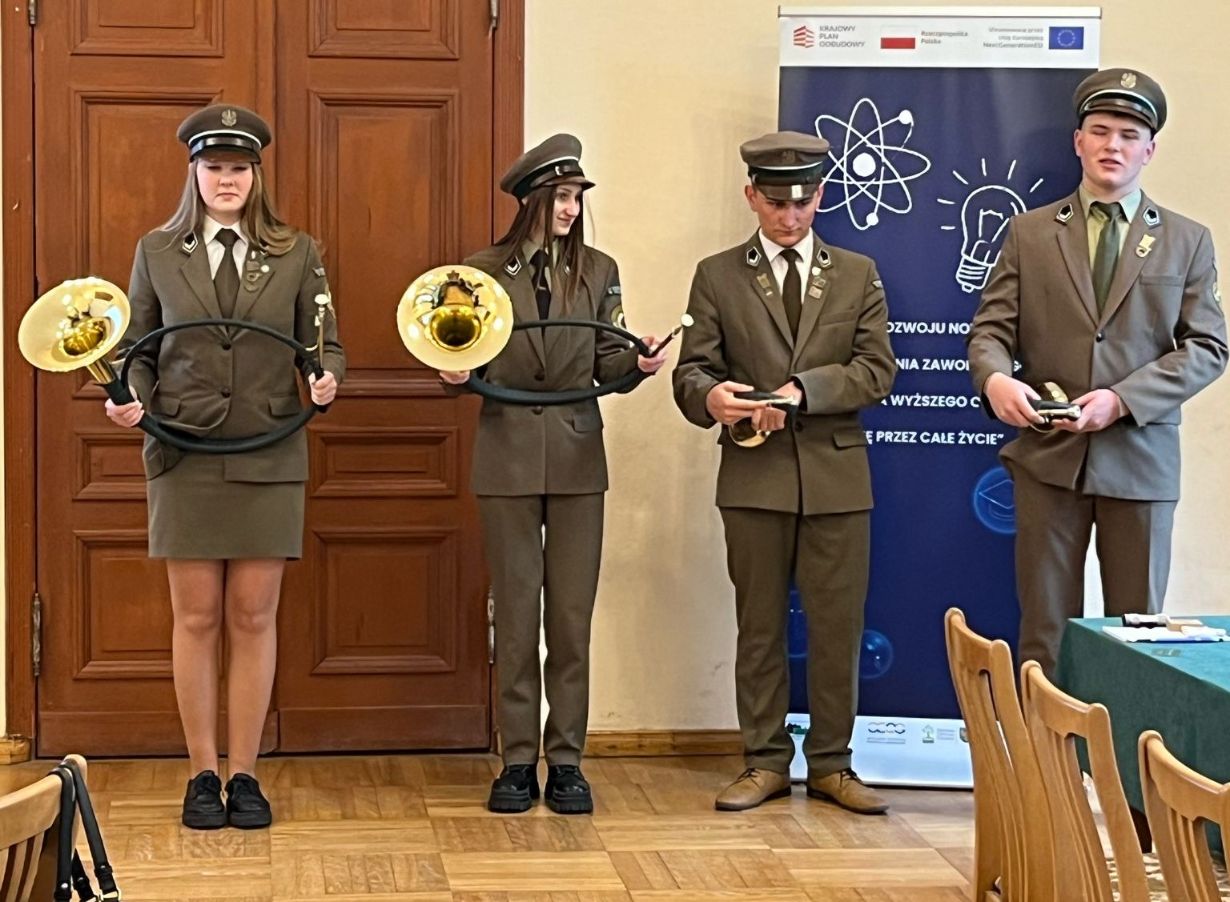 Czworo młodych ludzi w brązowych mundurach i czapkach stoi w pomieszczeniu. Dwie dziewczyny po lewej trzymają zakrzywione instrumenty dęte, a dwóch chłopców po prawej medale lub nagrody. Za nimi znajdują się drewniane drzwi i niebieski baner z grafiką o tematyce naukowej.