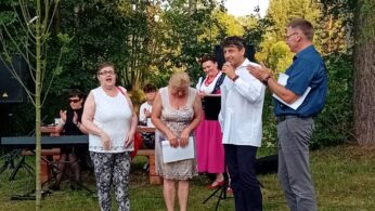 Grupa ludzi stoi na zewnątrz na trawie w pobliżu drzew. Trzy kobiety stoją z przodu, dwie się uśmiechają; jedna pochyla głowę. Dwóch mężczyzn stoi obok nich, jeden trzyma mikrofon i mówi. Inni siedzą i stoją w tle w pobliżu klawiatury. Atmosfera wydaje się świąteczna.