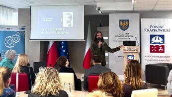 Mężczyzna z długimi włosami i brodą przemawia na podium w sali konferencyjnej. Za nim wyświetlany jest slajd ze zdjęciem i polskim tekstem, a także flagi, transparenty i siedzący widzowie, którzy uważnie słuchają.