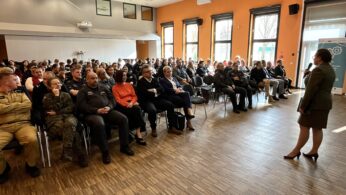 Kobieta w garniturze przemawia do dużej publiczności siedzącej w jasnym pomieszczeniu z pomarańczowymi ścianami i drewnianą podłogą; wśród uczestników są ludzie w mundurach i strojach biznesowych, z banerami i oknami widocznymi w tle.