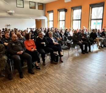 Kobieta w garniturze przemawia do dużej publiczności siedzącej w jasnym pomieszczeniu z pomarańczowymi ścianami i drewnianą podłogą; wśród uczestników są ludzie w mundurach i strojach biznesowych, z banerami i oknami widocznymi w tle.