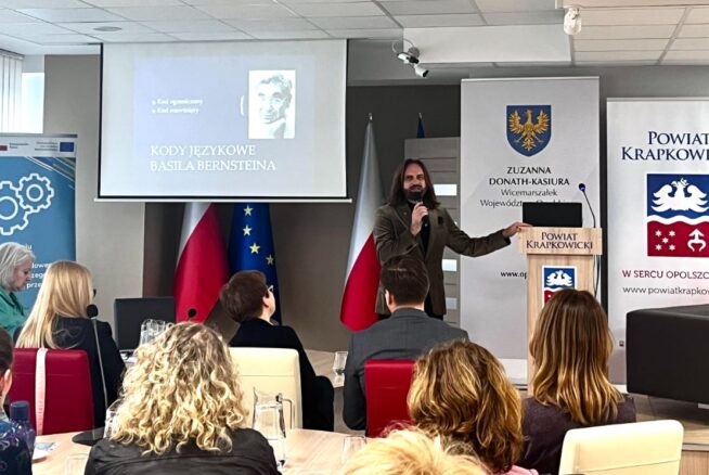 Mężczyzna z długimi włosami i brodą przemawia na podium w sali konferencyjnej. Za nim wyświetlany jest slajd ze zdjęciem i polskim tekstem, a także flagi, transparenty i siedzący widzowie, którzy uważnie słuchają.