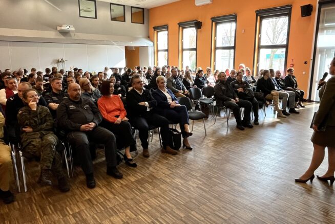 Kobieta w garniturze przemawia do dużej publiczności siedzącej w jasnym pomieszczeniu z pomarańczowymi ścianami i drewnianą podłogą; wśród uczestników są ludzie w mundurach i strojach biznesowych, z banerami i oknami widocznymi w tle.