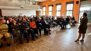 Kobieta w stroju biznesowym przemawia do siedzącej publiczności w dużym, jasnym pomieszczeniu z pomarańczowymi ścianami i drewnianymi podłogami. Zróżnicowana publiczność obejmuje ludzi w mundurach i zwykłych ubraniach, uważnie zwróconych w stronę mówcy. Okna wpuszczają światło dzienne z prawej strony.
