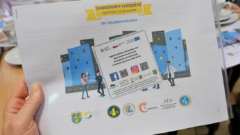 Ręka trzyma arkusz z kolorową grafiką przedstawiającą ludzi przed budynkami, różnymi logo, kodem QR i polskim tekstem o wydarzeniu tygodnia zawodowego w dniach 25-29 listopada 2024 roku. Na stole w tle widoczne są talerze i kubki.